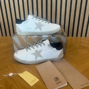 Golden Goose skor  - Snygga vita sneakers från Golden Goose med grå mockadetaljer och ikonisk stjärna på sidan. Skorna har grå snörning, svart hälparti och platt sula. Tillverkade i skinn och mocka för en lyxig känsla. Perfekta för dig som gillar stilrena och trendiga sneakers.