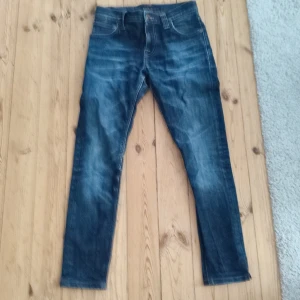Nudie Jeans blå slim fit jeans - Snygga blå jeans från Nudie Jeans med klassisk orange söm och vågig brodyr på bakfickorna. Jeansen har en slim fit passform, raka ben och är tillverkade i slitstarkt denim. Perfekta för dig som gillar en stilren och tidlös look. Jeansen är nedkortade så bör passa dig som är runt 155-165
