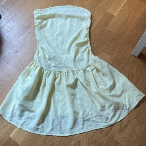 Ljusgul axelbandslös kortklänning - Söt ljusgul kortklänning med axelbandslös design och lätt rynkad kjol. Klänningen har en enkel och fräsch look, perfekt för varma dagar. Materialet känns mjukt och har en lätt struktur som ger en avslappnad vibe.