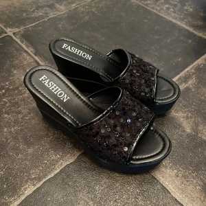 Svarta platåsandaler med paljetter - Svarta sandaler med hög platåsula och öppen tå. Ovandelen är dekorerad med svarta paljetter som ger extra glans. Snygg och trendig modell med bred rem och bekväm passform. Skorna kan användas på både ett festligt sätt och vanligt till dig som vill sticka ut lite extra.