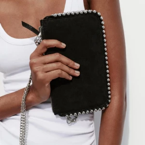 Svart mocka kuvertväska med kedja - Snygg svart kuvertväska i mocka med silverfärgade nitar längs kanterna och en lång kedjerem. Väskan har dragkedja upptill och en stilren, rektangulär form som ger en edgy vibe. Perfekt för att lyfta din outfit med lite extra attityd.