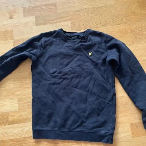Svart sweatshirt från Lyle & Scott - Svart sweatshirt från Lyle & Scott med klassisk rund hals och gul broderad logga på bröstet. Tröjan har långa ärmar och är tillverkad i mjuk bomull, perfekt för en chill och stilren look.