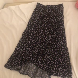 Blommig långkjol från Lindex - Supersöt långkjol från Lindex med svart botten och små lila blommor över hela kjolen. Kjolen har en lätt utsvängd form och är perfekt för dig som gillar en romantisk och chill stil. Materialet känns mjukt och flowy, perfekt för vår och sommar.