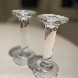 2 ljusstakar i glas - Bilderna visar två eleganta ljusstakar i glas, perfekta för att skapa mysig stämning hemma. De har en stilren design som passar både till vardag och fest. Aldrig använda 
