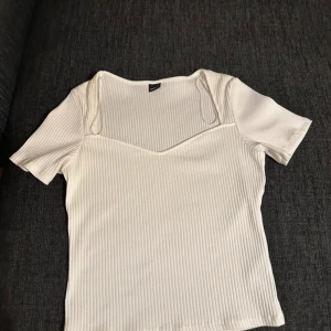 Vit ribbad topp med v-ringning - Snygg vit ribbad topp med korta ärmar och v-ringad hals. Toppen har en figurnära passform och är perfekt att styla med jeans eller kjol. Materialet är mjukt och stretchigt för extra komfort. Aldrig använd 