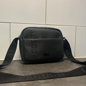 Svart axelväska/midjeväska från Lacoste - Säljer en snygg svart axelväska från Lacoste!  justerbar axelrem vilket gör att den funkar minst lika bra som en midjeväska. Det kan du se på näst sista bilden. Väskan har 4 olika fack vilket gör att den kan hålla mycket utrymme. dragkedjorna på väskan är helt felfria och funkar perfekt. Stilren och funkar till många olika outfits och stilar. Om du vill veta mer eller har fler frågor så tveka inte på att skriva!