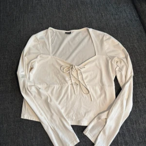 Vit långärmad topp med snörning - Snygg vit långärmad topp med fyrkantig urringning och snörning framtill. Croppad modell som passar perfekt till jeans eller kjol. Tunn och mjuk i materialet, superenkel att styla för en trendig look.