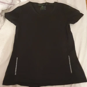 Svart mesh t-shirt med reflexdetaljer - Svart t-shirtmed korta ärmar och meshpaneler på sidorna och ärmarna för extra ventilation. Snygga reflexdetaljer längs sidorna ger en sportig vibe. Perfekt för dig som gillar en enkel men cool stil.