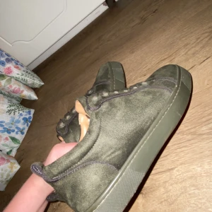 Olivgröna sneakers från Louboutin - Snygga olivgröna sneakers från Christian Louboutin i mocka med klassisk röd sula och diskreta sömmar. Skorna har snörning och en platt sula, perfekt för dig som vill sticka ut med exklusiva detaljer och streetkänsla.