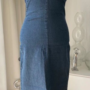 Axelbandslös jeansklänning i blå denim - Snygg axelbandslös långklänning i blå denim med figurnära passform. Klänningen har en slits bak för extra rörelsefrihet och markerade sömmar som ger en cool look. Perfekt för dig som vill sticka ut med en modern och trendig jeansstil.
