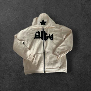 over sized Beige hoodie med svart stjärna och tryck - Snygg beige hoodie med svart stjärna på huvan och svart broderat tryck framtill. Tröjan har dragkedja hela vägen, fickor på sidorna och långa ärmar. Perfekt för dig som gillar streetwear och vill sticka ut med coola detaljer.