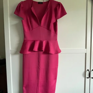 Rosa peplumklänning från Club L - Snygg rosa långklänning från Club L med peplumdetalj i midjan och v-ringad hals. Klänningen har korta ärmar och figurnära passform som framhäver siluetten. Perfekt för dig som vill sticka ut med en färgstark look.