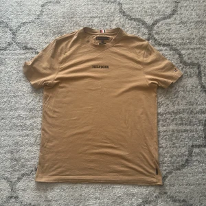 Beige t-shirt från Tommy Hilfiger - Snygg beige t-shirt från Tommy Hilfiger med mörkblått 'HILFIGER'-tryck på bröstet. Klassisk rund halsringning och korta ärmar. Tillverkad i mjuk bomull och har en regular fit. Perfekt för en clean och stilren look.