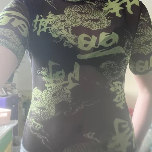Svart mesh topp med drakmönster Missguided - Säljer en svart mesh topp från Missguided med coolt limegrönt drakmönster och asiatiska tecken. Toppen är tight och har korta ärmar, perfekt för dig som vill sticka ut. Materialet är genomskinligt och stretchigt syntet. Köpt från Sellpy