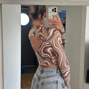 Brun mönstrad mesh-top från Amisu - Säljer en croppad långärmad topp från Amisu med swirl-mönster i bruna och beige toner. Toppen har öppen rygg och är tillverkad i tunn, stretchig mesh som sitter tight. Tröjan går att använda med öppningen antingen  fram eller bak. 