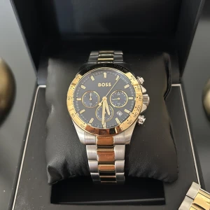 BOSS klocka i guld och silver - Stilren klocka från BOSS med blå urtavla, guldiga detaljer och kronograf. Armbandet är i rostfritt stål med både silver- och guldfärgade länkar. Klockan har tre små urtavlor, datumvisning och markerade index. Perfekt accessoar för dig som gillar en lyxig look. Nypris 3595