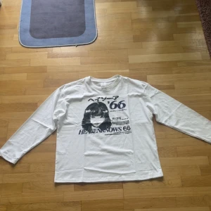 Vit långärmad tröja med anime-tryck - Vit långärmad tröja med svart anime-inspirerat tryck och japansk text på bröstet. Tröjan har en rak passform och är gjord i mjuk bomull, perfekt för dig som gillar streetstyle och grafiska prints.