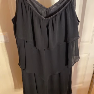 Svart omlottklänning med volang - Svart klänning med tunna axelband och v-ringning. Klänningen har dubbla lager av tunn, luftig polyester och volangdetaljer framtill. Den är ärmlös och har en omlottliknande design som ger ett flowy intryck.