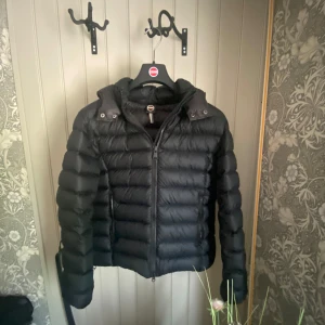Svart Colmar dunjacka  - Säljer en svart dunjacka från Colmar med avtagbar huva och dragkedja framtill. Jackan har en clean look med diskret logga på ärmen och är perfekt för kalla dagar. Fylld med dun och har en klassisk pufferstil som är både snygg och bekväm.