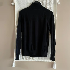 Svart polotröja från Bläck - Klassisk svart polotröja från Bläck med ribbad hög krage och långa ärmar. Tröjan är i mjuk bomull och har en stilren, enkel design som funkar till allt. Perfekt för lager-på-lager och snygg till både jeans och kostymbyxor.