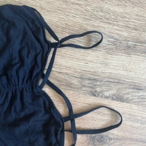 Svart bandeau topp med smala band - Säljer en svart bandeau topp med smala axelband och snygg rynkning framtill. Toppen har en croppad passform och dubbla band som korsas framtill för en cool detalj. Perfekt att styla med höga jeans eller kjol.