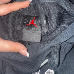 Svart Jordan hoodie med tryck - Svart hoodie från Jordan med stort grafiskt tryck och Jumpman-logga på bröstet samt ärmen. Klassisk känguruficka och snörning i huvan. Perfekt för dig som gillar streetwear och sportig stil.