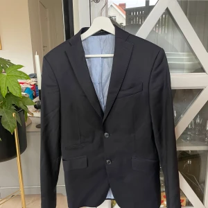 Svart kavaj från Zara Man, strl 46 - Stilren svart kavaj från Zara Man i storlek 46. Klassisk modell med två knappar framtill, fickor med lock och en bröstficka. Fodret är ljusblått med tunna ränder. Perfekt för en dressad look.