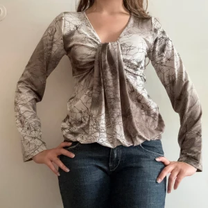 Beige omlottopp med v-ringning - Säljer en beige omlottopp med abstrakt mönster i bruna toner. Toppen har v-ringning, långa ärmar och snygga draperingar framtill som ger en cool och unik look. Materialet är glansigt och känns mjukt mot huden. Perfekt för dig som gillar statement-plagg!