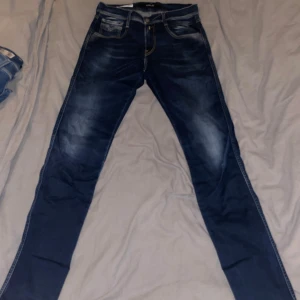 Replay jeans Anbass mörkblå - Snygga mörkblå jeans från Replay med klassiska fem fickor. Jeansen har slitningar och ljusare partier framtill för en cool, använd look. Tydliga kontrastsömmar och läderpatch bak i midjan. 