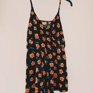 Blommig jumsuit / shortsdress från H&M Divided - Supersöt svart shortsdress med orange blommor från H&M Divided. Klänningen har smala axelband, omlott-v-ringning och ett flowy fall. Perfekt för sommarens alla tillfällen och lätt att styla med sneakers eller sandaler.  Helt & fint skick dock små tvättludd