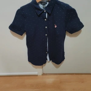 Mörkblå prickig skjorta U.S. Polo Assn. - Mörkblå skjorta från U.S. Polo Assn. med små vita prickar och klassisk krage. Skjortan har korta ärmar, knäppning framtill och en broderad rosa logga på bröstet. Tillverkad i bomull med lite stretch för extra komfort. Passar både tjejer och killar 