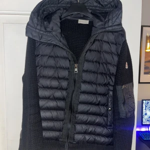 Svart dunjacka från Moncler med huva - Säljer en svart Moncler dunjacka med stickade ärmar och quiltad front. Jackan har huva, dragkedja framtill och en snygg Moncler-logga på ärmen. Perfekt för dig som vill ha en stilren och varm jacka med exklusiv känsla.