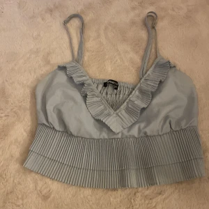 Ljusblå volangtopp från Gina Tricot - Supersöt ljusblå linnetopp från Gina Tricot med smala axelband, v-ringning och volangdetaljer både upptill och nertill. Toppen har plisserade volanger och resår i ryggen för en bekväm passform. Perfekt för sommarens alla tillfällen!