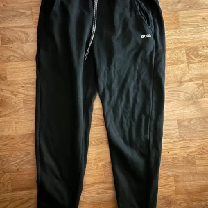 Svarta mjukisbyxor från BOSS - Svarta mjukisbyxor från BOSS med broderad logga på vänster ficka. Byxorna har elastisk midja med snörning och två sidofickor. Tillverkade i mjukt bomullsmaterial, perfekta för chill eller träning.