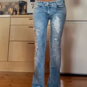 Ljusblå slitna bootcut jeans - Säljer ett par ljusblå jeans med slitna detaljer och hål på låren. Jeansen har bootcut-modell med lätt utsvängda ben och klassisk femficksdesign. Perfekta för dig som gillar en avslappnad och trendig look.