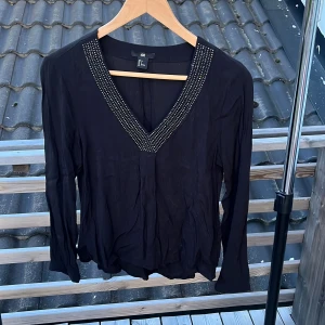 Svart blus med v-ringning från H&M - Svart blus från H&M i storlek 34 med lång ärm och snygg v-ringning dekorerad med små silvriga nitar. Blusen har en lös passform och är tillverkad i ett lätt, mjukt tyg som ger en skön känsla. Perfekt för dig som gillar stilrena detaljer.