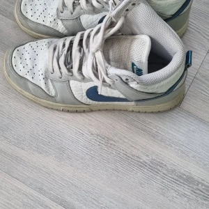 Nike Dunk Low vit/grå/blå 36.5 - Nike Dunk Low sneakers i vitt och ljusgrått skinn med blå swoosh. Klassisk siluett med perforerad tå, snörning och låg profil. Bekväm mesh på insidan och slitstark yttersula. Perfekta för dig som gillar streetstyle och vill sticka ut med en ikonisk modell.