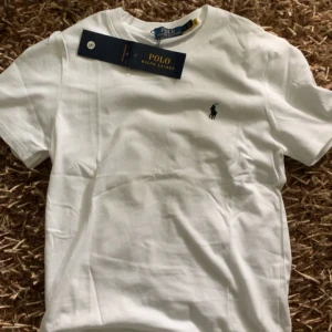 Vit t-shirt från Polo Ralph Lauren - Vit klassisk t-shirt från Polo Ralph Lauren med broderad svart logga på bröstet. T-shirten är kortärmad och har en rund halsringning. Tillverkad i mjuk bomull för en skön och stilren look. Perfekt till jeans eller shorts.