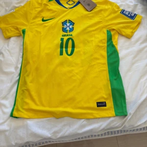 Brasilien Neymar Jr 10 Nike fotbollströja - Gul Brasilien fotbollströja från Nike med gröna sidopaneler och blå detaljer. Nummer 10 och Neymar Jr tryckt på ryggen, CBF-emblem på bröstet och Dri-FIT-material som andas. Klassisk passform med korta ärmar och rund hals.