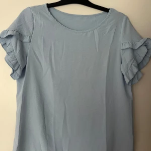 Ljusblå blus med volangärmar - Supersöt ljusblå blus med rund halsringning och korta ärmar med dubbla volangdetaljer. Perfekt för dig som gillar en feminin och luftig stil. Tillverkad i mjuk bomull som känns skön mot huden.