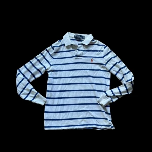 Ralph Lauren piké  - Vit och blå randig långärmad pikétröja från Polo Ralph Lauren. Klassisk krage, knäppning vid halsen och den ikoniska röda loggan broderad på bröstet. Tillverkad i bomull för en skön och avslappnad känsla. Har några gulafläckar som bör lossna med blekningsmedel syns på sista bilden ! 