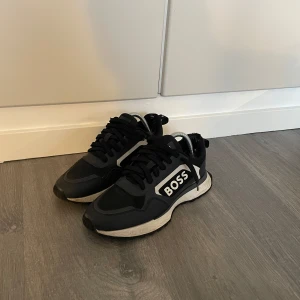  BOSS skor - Snygga svarta sneakers från BOSS med vit logga på sidan och detaljer i vitt och grått. Skorna har snörning och en chunky vit sula. Tillverkade i en mix av syntet och textil för en sportig look. Perfekta för dig som gillar stilrena och trendiga sneakers.