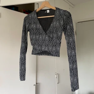 Svart omlottopp med glitter - Snygg svart omlottopp med silvrigt glittrigt geometriskt mönster. Toppen är croppad, har lång ärm och djup v-ringning. Perfekt för dig som vill sticka ut med coola detaljer och en trendig look.