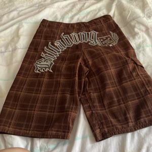 Bruna rutiga Billabong shorts - Snygga bruna shorts från Billabong med rutigt mönster och stora loggan på baksidan. De har svart snörning i midjan och en liten ficka med broderad detalj i rött, gult och grönt. Perfekta för sommaren och strandhäng!