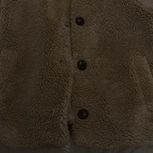 Beige teddyjacka från ONLY - Supermjuk och fluffig teddyjacka från ONLY i beige färg. Jackan har stora knappar framtill, klassisk krage och två fickor. Perfekt för dig som vill ha en mysig och trendig look under kyliga dagar.