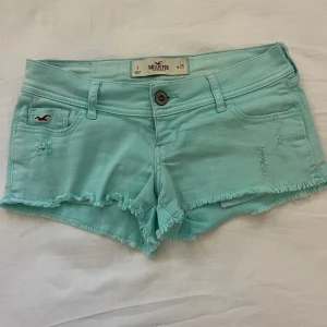 Turkosa jeansshorts från Hollister - Snygga turkosa jeansshorts från Hollister med fransig kant och slitna detaljer. Klassisk femficksmodell med låg midja och knappstängning. Perfekta för sommardagar och festivalhäng. Ikonisk Hollister-logga på fickan och bak. Jätte lågmidjade! 💕