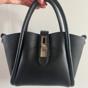 Svart handväska i skinn med metallspänne - Snygg svart handväska i präglat skinn med två rejäla handtag och ett stilrent silverfärgat metallspänne framtill. Ifrån Gina tricot