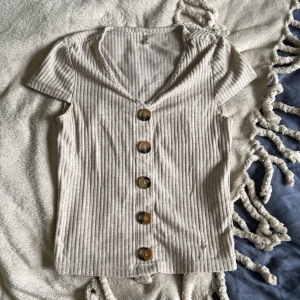 Beige ribbad topp från ONLY - Säljer en beige ribbad topp från ONLY med korta ärmar och v-ringning. Toppen har stora bruna knappar framtill. Knappt använd