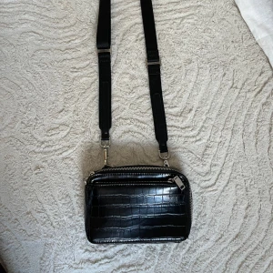 Svart axelväska från Zara - Säljer en svart axelväska från Zara med croco-mönstrad yta och bred justerbar rem. Väskan har två stora fack med silvriga dragkedjor och en stilren, boxig form. Perfekt för dig som vill ha en snygg och praktisk väska.