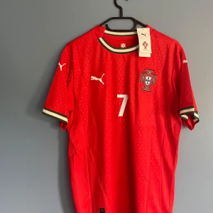 Portugal Ronaldo 7 fotbollströja Puma - Riktigt snygg Portugal fotbollströja från Puma med Ronaldo och nummer 7 på ryggen. Tröjan är röd med vita detaljer, broderat landslagsmärke och subtilt mönster i tyget. Kortärmad modell med ribbade muddar i grönt, rött och vitt. Perfekt för dig som älskar fotboll och Ronaldo.